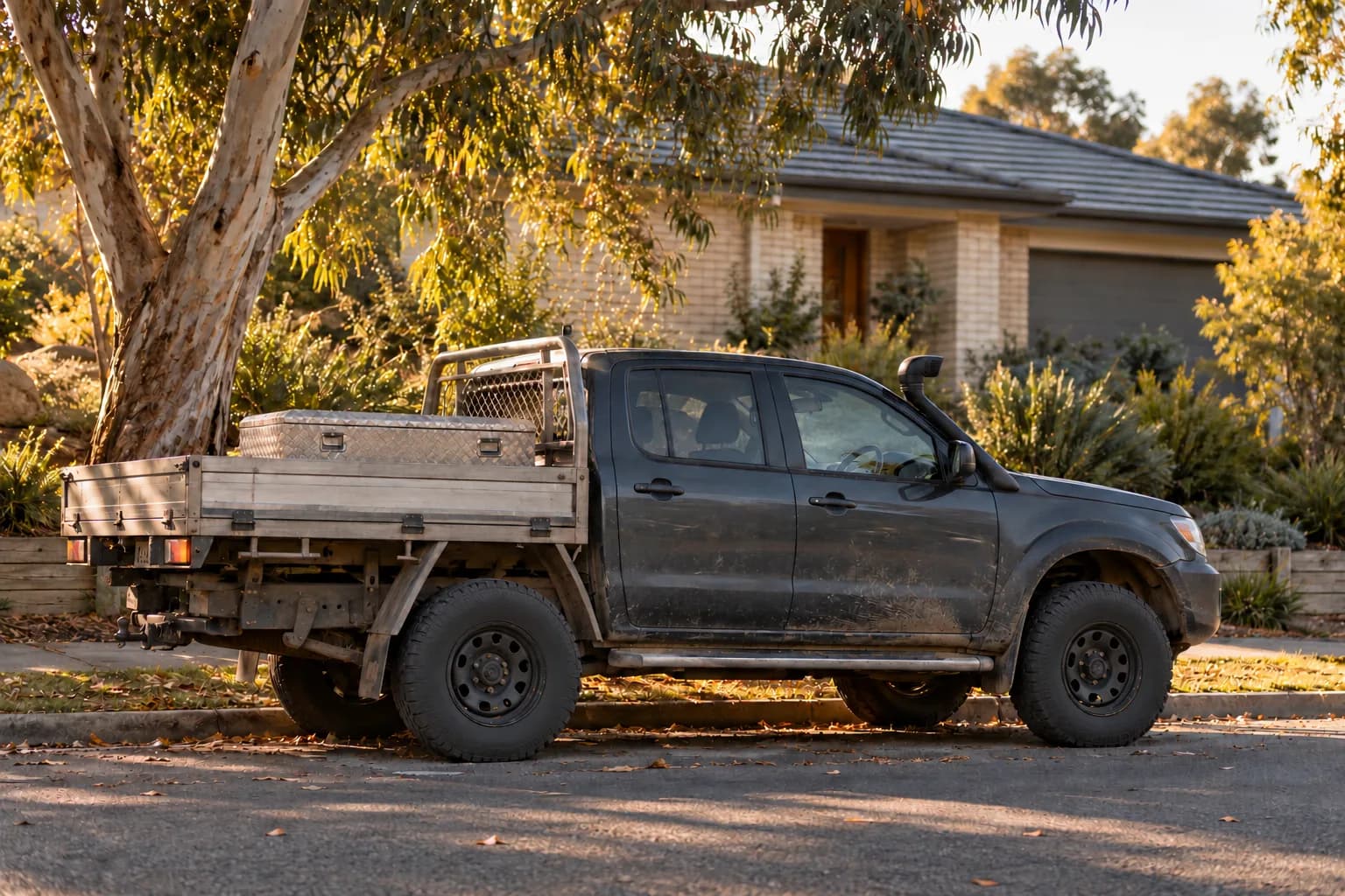 A dusty ute with a toolbox is parked outside a suburban brick home in afternoon light.