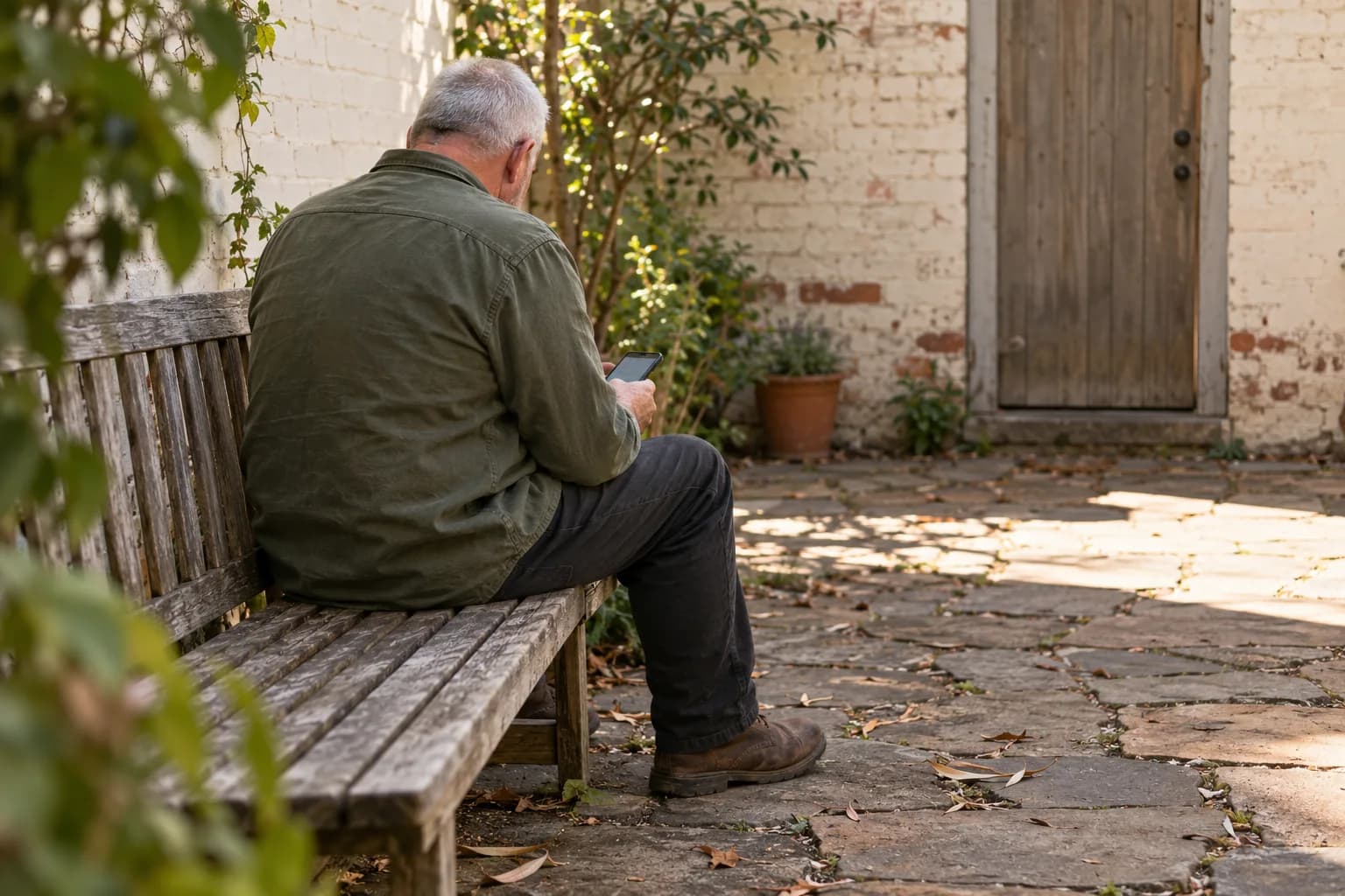 An older man checks his phone on a timber bench in a quiet brick courtyard.